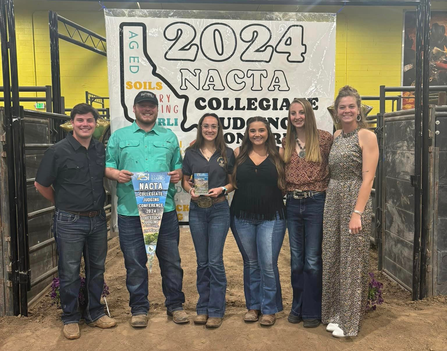 Two male students and four female students, 2 of which are holding NACTA awards, stand in front of an empty animal pen with a NACTA banner hanging behind them.